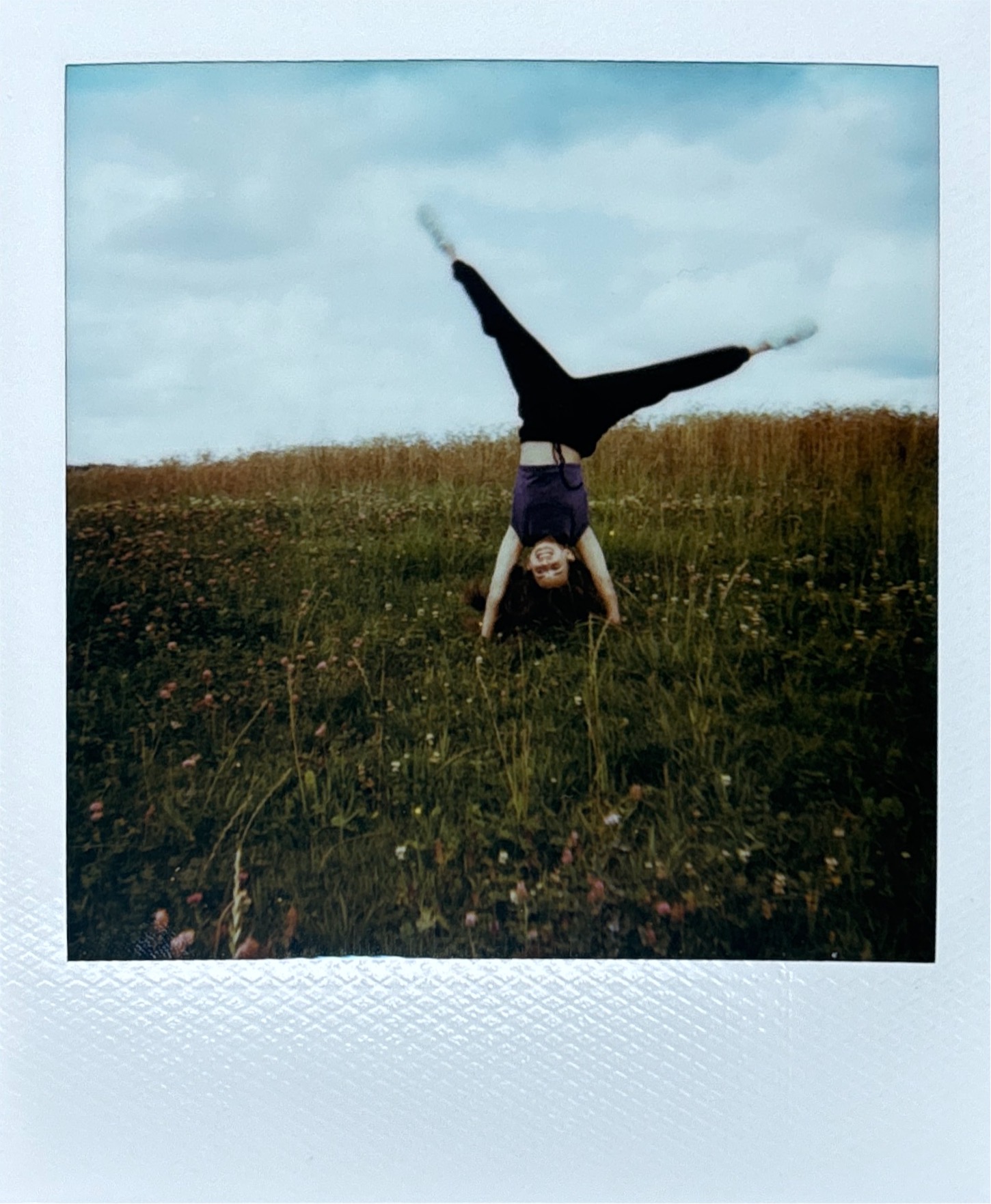 Polaroid snapshot of a person doing a handstand in a grassy field. The subject is silhouetted against an overcast sky, wearing dark clothing with legs spread in a V-shape. Wild grass and small flowers are visible in the foreground, captured with the soft, muted colors characteristic of vintage instant photography.
