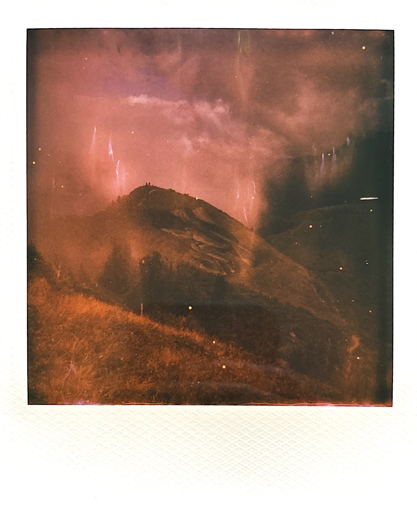 Heavily saturated and overexposed Polaroid photograph dominated by warm red and orange tones. The image appears to show a mountainous landscape or rock formation, but is heavily affected by light leaks and color shifts, creating an abstract, dreamlike quality typical of aged or heat-damaged instant film.