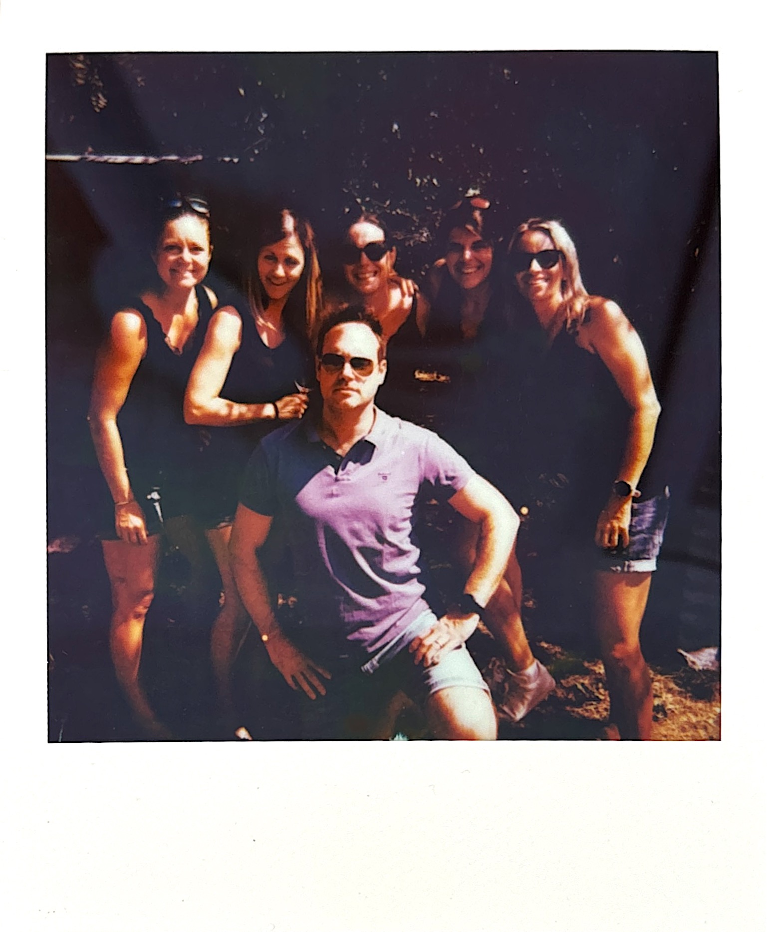 Polaroid group portrait of six friends posing outdoors on a sunny day. Five women stand in the back row wearing summer clothes and sunglasses, while one person in a light purple shirt and sunglasses sits in front. The photo has a warm, nostalgic quality with natural lighting and the classic instant film aesthetic.