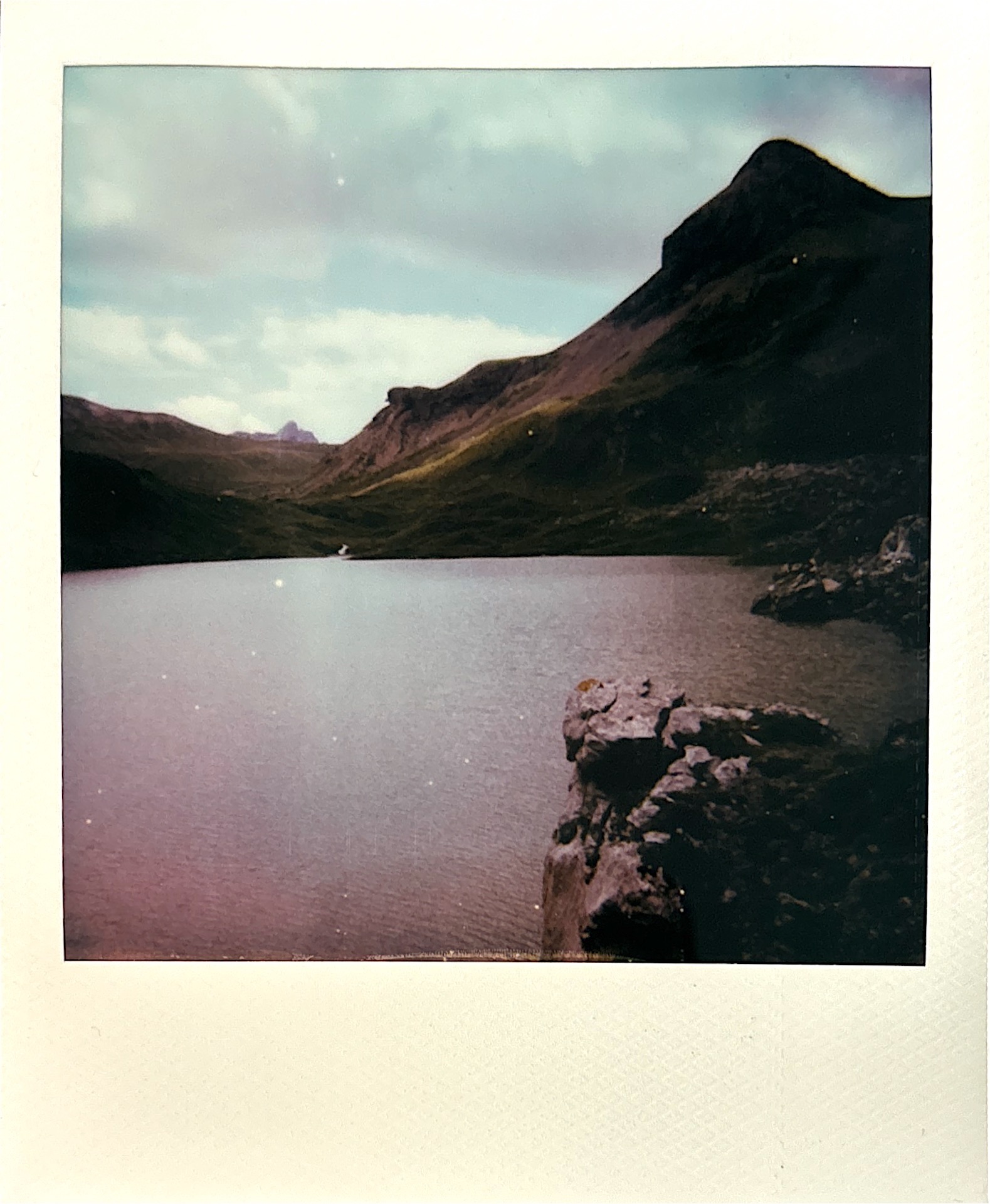 A serene view of Sanetsch Lake, with calm waters reflecting the surrounding mountainous terrain under a partly cloudy sky.