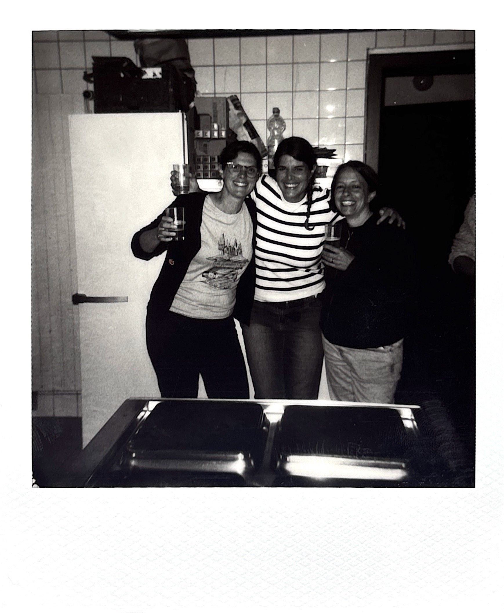 A black and white image of three people smiling and posing together in a kitchen, each holding a drink and embracing each other.