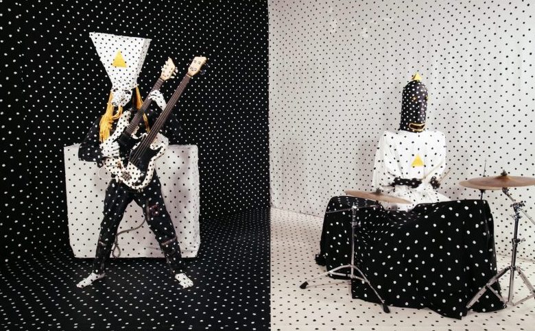 This image depicts two members of the band "Angine de Poitrine." They are dressed in striking, patterned outfits against a matching polka dot background. The band member on the left is energetically playing a bass guitar with a high, angular headdress, while the member on the right is seated at a drum set, with their face obscured by a hood. The scene is visually bold and artistic, showcasing the band's unique and theatrical style.