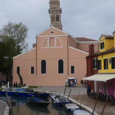 Church of San Martino in Burano with its leaning campanile, colourful houses nearby and boats moored along the canal in front