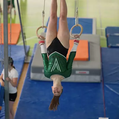Female gymnast in green Gym Ursy leotard hanging upside down on gymnastics rings, legs extended straight up