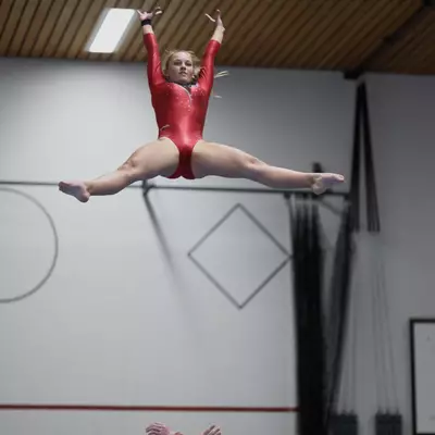 Female gymnast in red leotard thrown into the air by male partners, performing a straddle split jump high above the floor