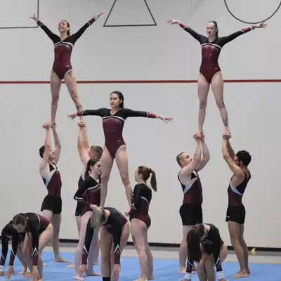 Group of gymnasts in dark red and black leotards forming a multi-level human pyramid during a group acrobatics routine