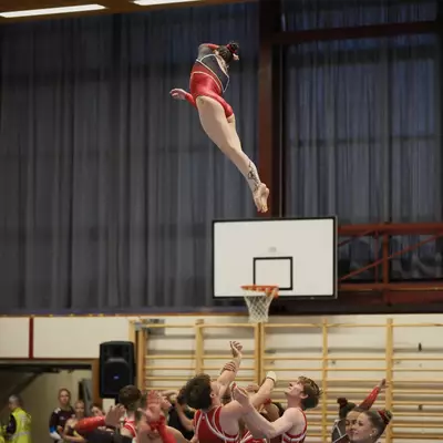 Female gymnast in red leotard launched high into the air by a group of teammates during an acrobatic group routine