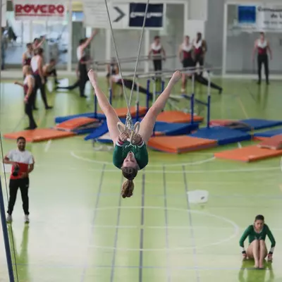 Female gymnast in green leotard hanging upside down on rings, wide shot of the gymnasium with male gymnasts warming up on parallel bars in the background