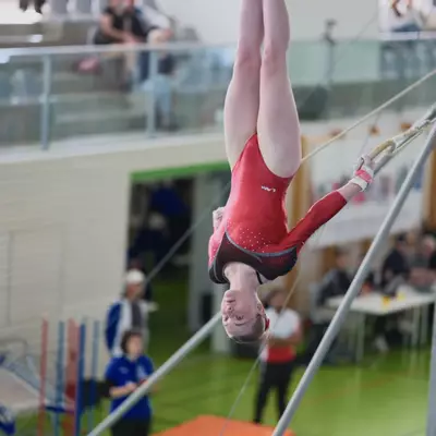 Female gymnast in red leotard hanging upside down on rings during the official competition, spectators visible in the stands behind her