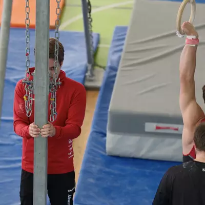 A coach in a red jacket adjusts the rings height mechanism while a male gymnast holds the rings with arms raised