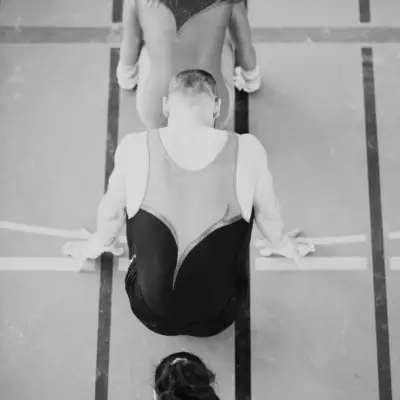 Black and white overhead shot of three gymnasts aligned on a gymnastics mat, seen from behind, waiting their turn
