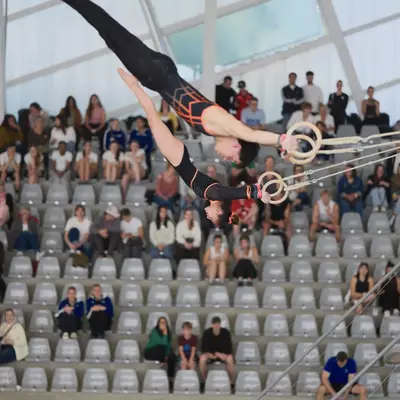Gymnast in black and orange leotard performing a dramatic horizontal back lever position on rings, audience packed in the bleachers behind