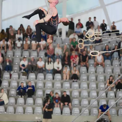 Male gymnast in black and orange leotard performing an inverted tuck on rings, audience in the bleachers behind him