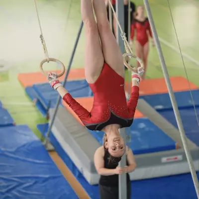 Female gymnast in red leotard hanging upside down on rings, smiling, with a teammate spotting her from below