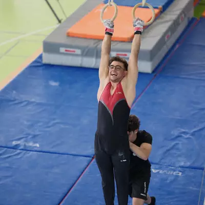 Male gymnast in red leotard laughing broadly while hanging on rings, a teammate helping spot him from below