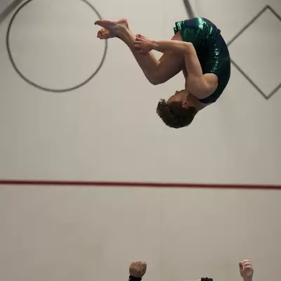 Male gymnast in green iridescent leotard performing a tucked back somersault in mid-air after being thrown by partners