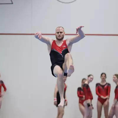 Male gymnast in red leotard leaping with one knee raised during a floor exercise, teammates in red watching from the side