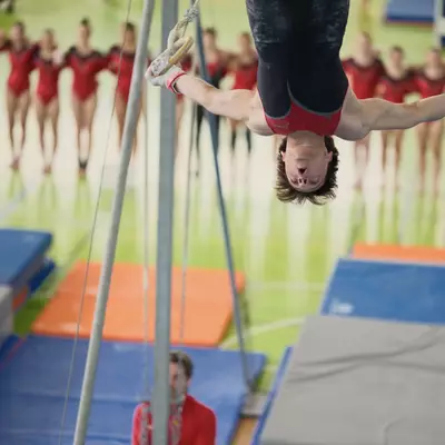 Male gymnast in red and black leotard performing an upside-down position on rings, the full team in red lined up watching in the background