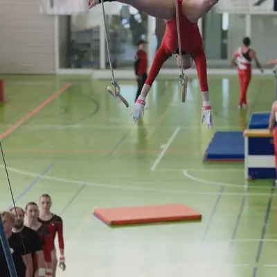Female gymnast in red leotard performing an upside-down straddle split on rings, teammates waiting in line watching from the side