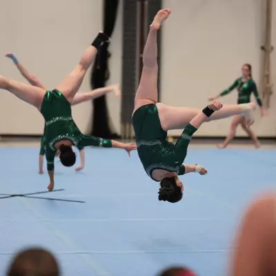 Two female gymnasts in green leotards simultaneously performing back handsprings on the floor exercise mat