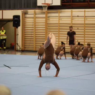 Gymnast in brown leotard performing a handstand at the center of the floor, teammates crouched in synchronized positions around her
