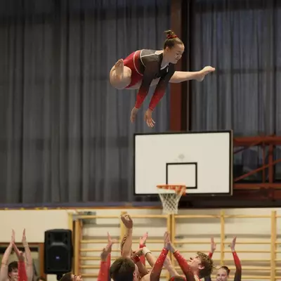 Female gymnast in red and black leotard thrown high into the air by teammates in a flying layout, arms reaching upward