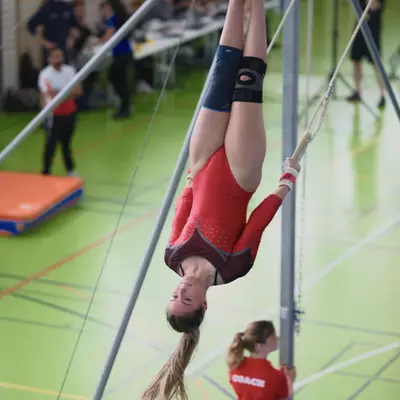 Female gymnast in red leotard hanging upside down on rings during competition, a coach in red jacket visible on the floor below, spectators in the background