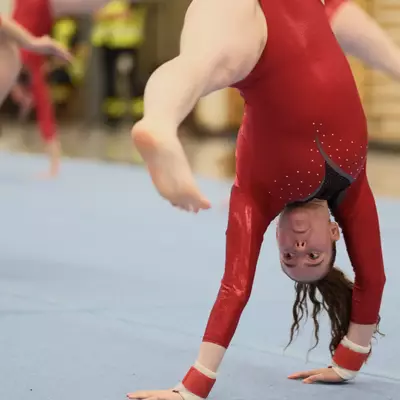Female gymnast in red leotard performing a one-handed cartwheel on the floor, close-up showing her inverted face and flying hair