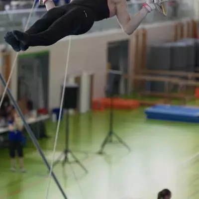 Male gymnast in red and black leotard performing a swallow position high on the rings, a female gymnast doing floor exercise visible far below