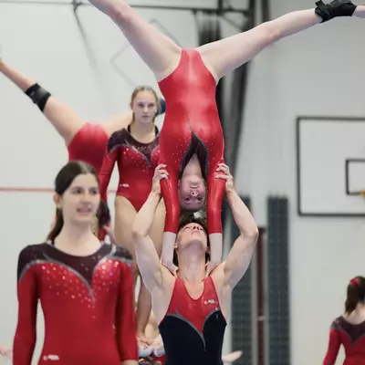 Acrobatic pairs lift: a male gymnast holds a female gymnast upside down overhead at arm's length during a group routine
