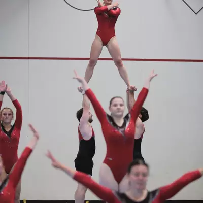 Female gymnast in red leotard elevated overhead by two male gymnasts in a pyramid lift during a group acrobatic routine