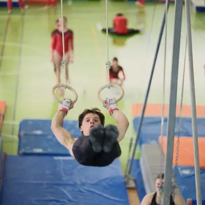 Male gymnast gripping the rings and looking upward with intensity, other gymnasts warming up in the background