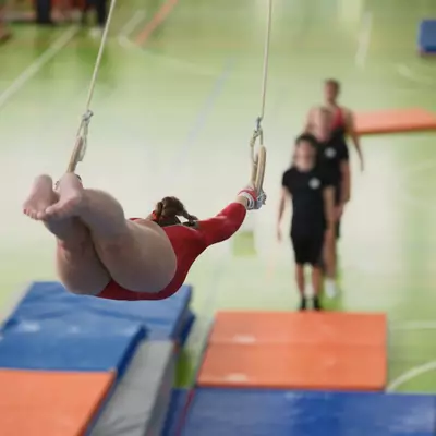 Female gymnast in red leotard swinging on rings with legs tucked, coaches and spectators visible in the blurred background