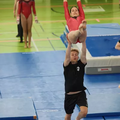 A female gymnast in a red leotard hanging from gymnastic rings while a male spotter in black lifts her legs from below, with other gymnasts in red visible in the background.