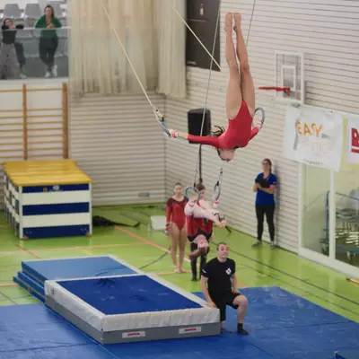 Wide competition view of a female gymnast in a red leotard performing an inverted position on rings at center, with another gymnast also inverted on rings in the background, coaches, and spectators across the sports hall.