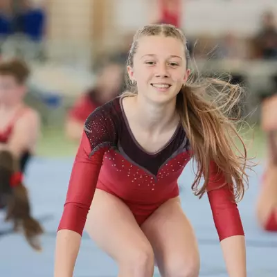 Close portrait of a smiling young female gymnast in a red leotard raising both arms joyfully, with another gymnast performing a back bend partially visible beside her and a packed audience in the background.