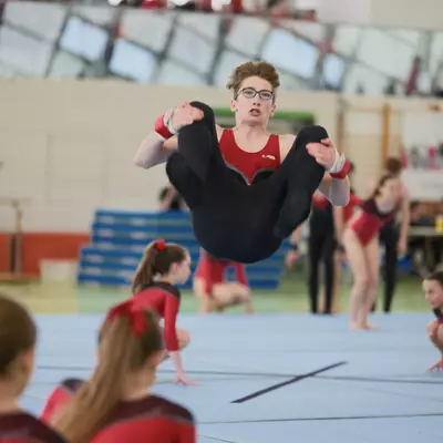 A male gymnast wearing glasses in a red and black outfit performing a high tucked jump above the floor mat, knees drawn toward his hands with an intense expression, surrounded by crouching gymnasts in red.