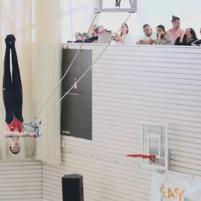 A gymnast in a black and red outfit performing a full inverted body hold on gymnastic rings high above the competition floor, with spectators leaning over a railing above and sponsor banners visible below.