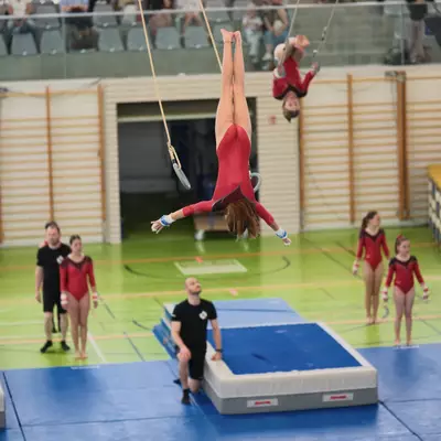 Three gymnasts in red leotards simultaneously performing inverted positions on gymnastic rings in a sports hall, with coaches in black watching from below and spectators visible in the upper gallery.